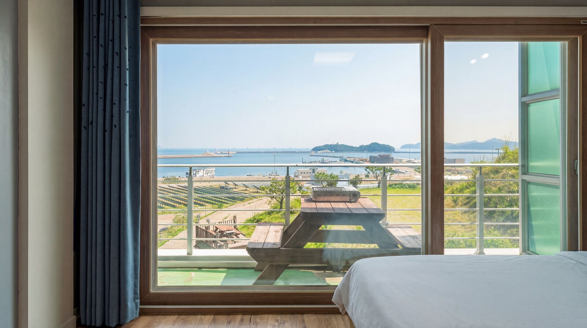 A bedroom with a large window overlooking a terrace with a view of the sea and distant land.