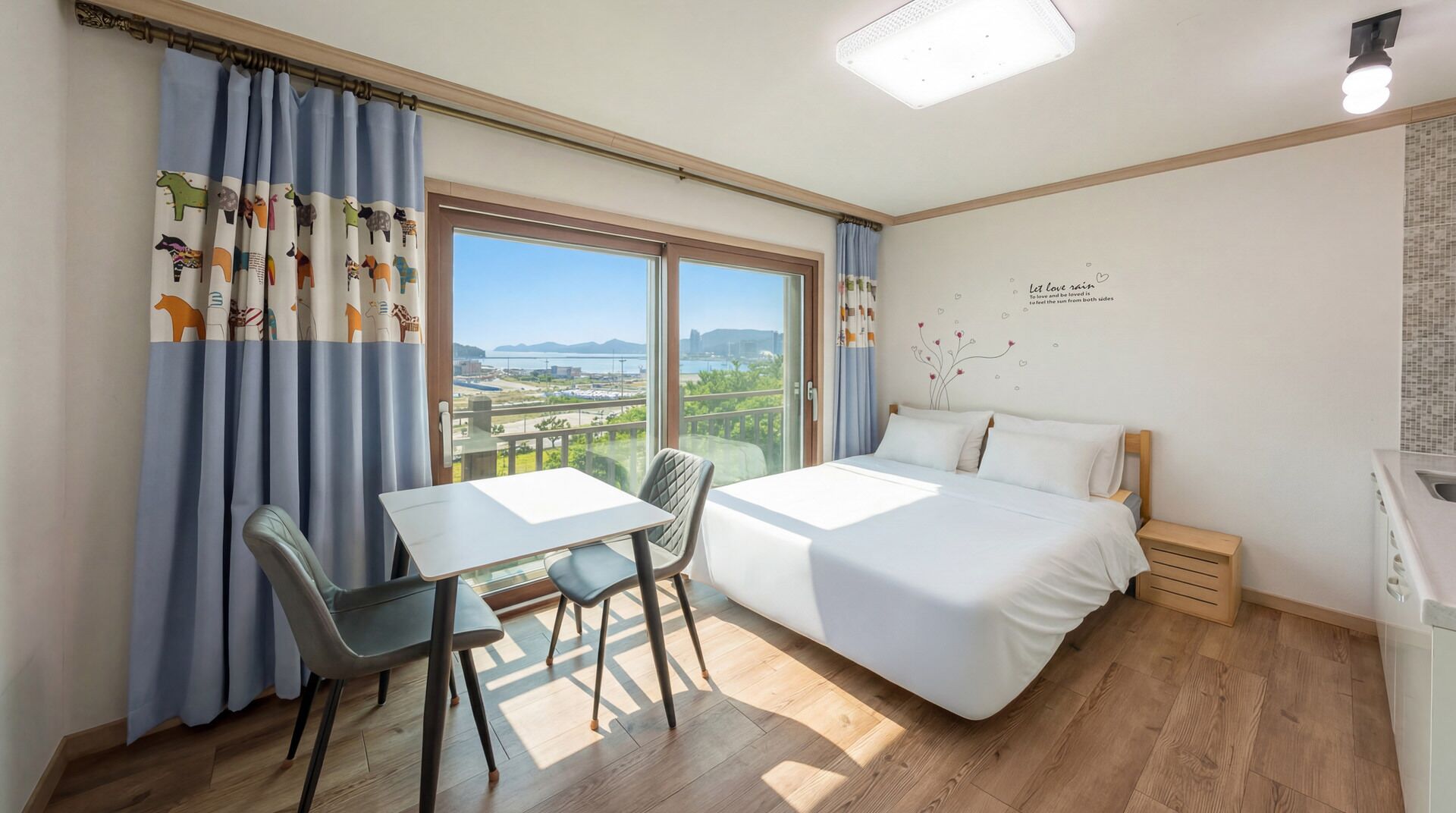 Quarto de hotel com cama, mesa, duas cadeiras, varanda e vista, além de parede com frase decorativa.