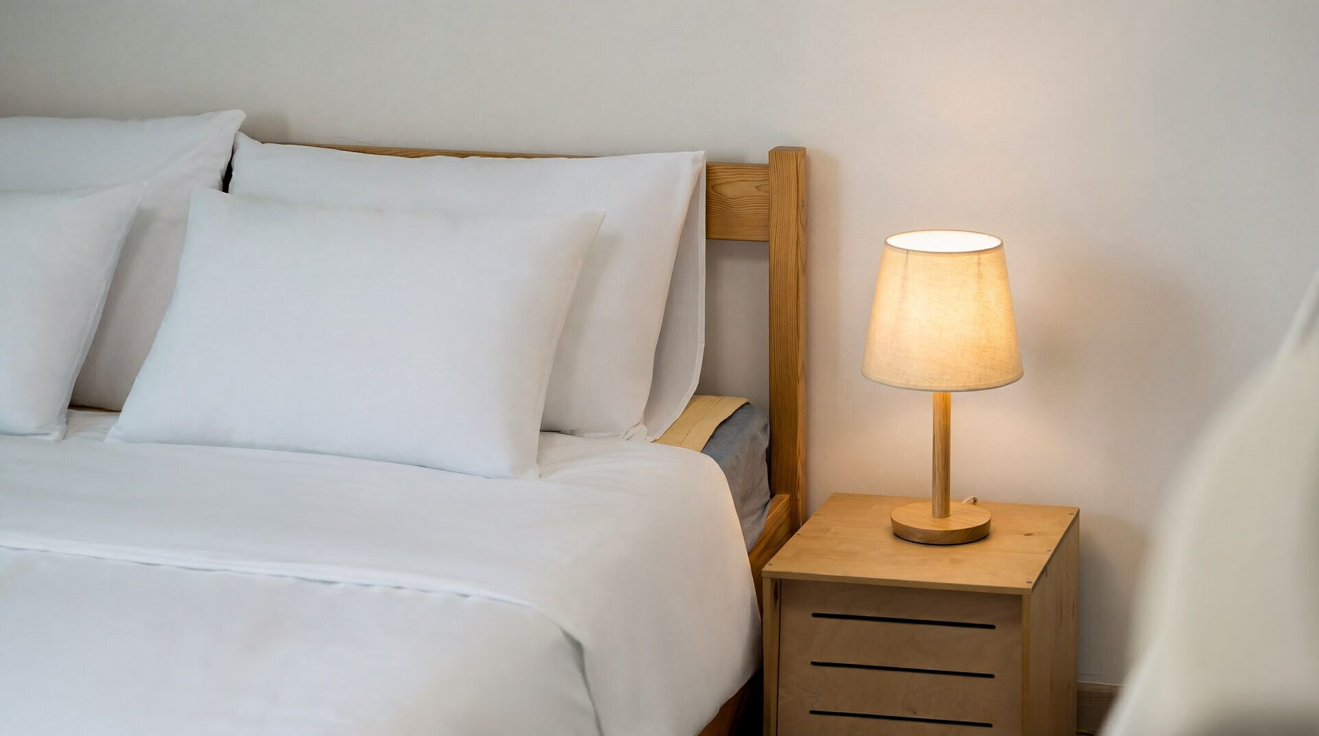 A neatly made bed with white linens and pillows, a wooden bedside table with a lamp, and a wooden nightstand with drawers.