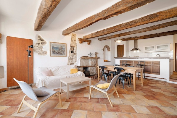 A living room with a sofa, chairs, a table, and a kitchen area with a tiled backsplash and wooden beams.