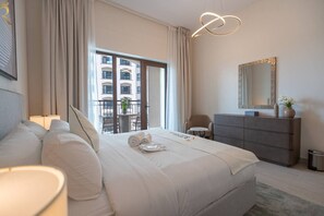 Une chambre d’hôtel avec un grand lit, une commode, une chaise, un balcon donnant sur des immeubles et un luminaire moderne.