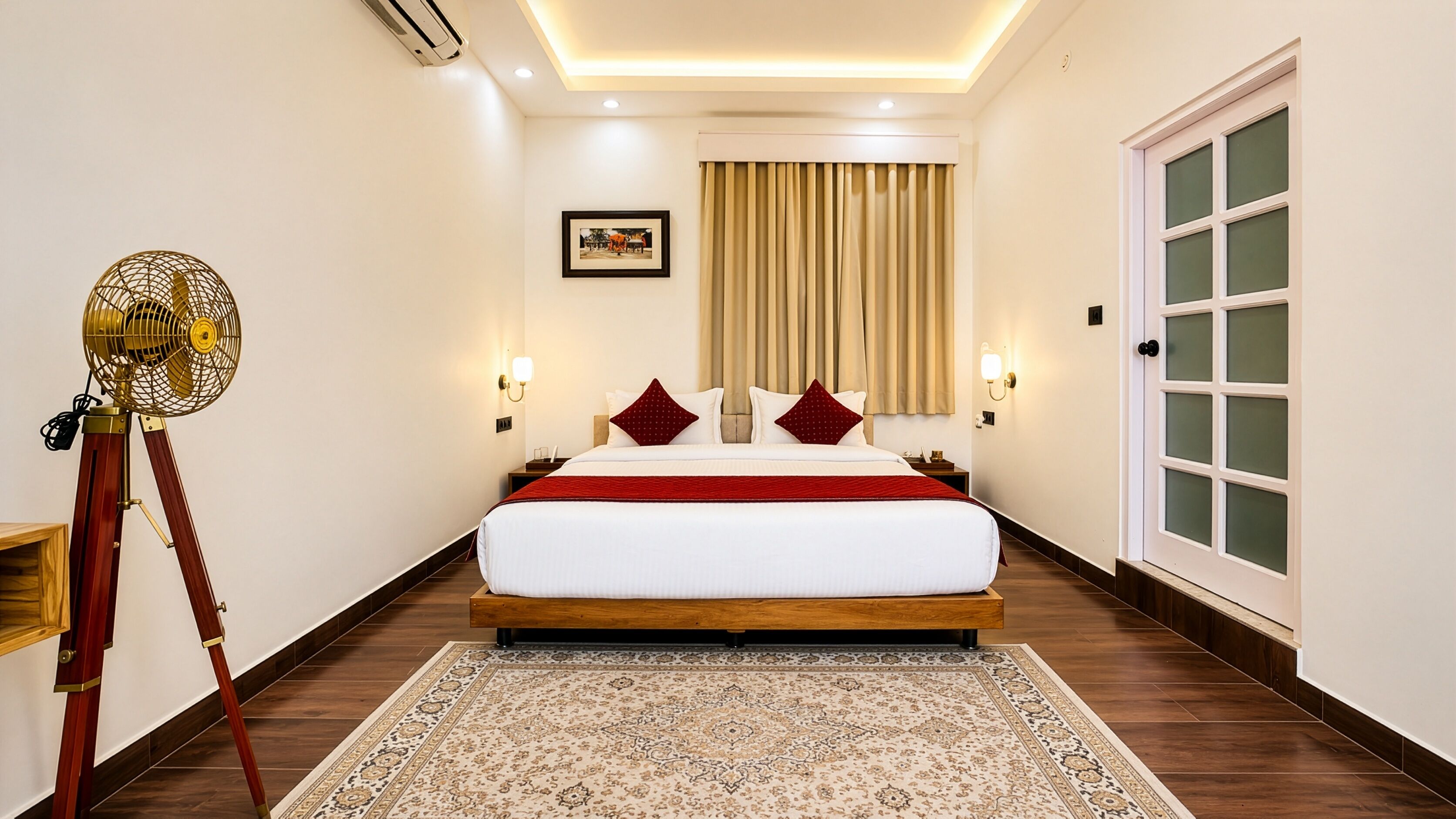 A hotel room with a large bed, a standing fan, a framed picture on the wall, and a door with frosted glass panels.
