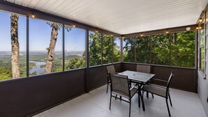 A covered outdoor area with a table and chairs, large windows offering a view of trees and a body of water. - Cabin at the Gap | You Won't Believe the View! (Harpers Ferry)