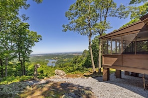 A wooden cabin with large windows, surrounded by trees and a view of a lake and valley.