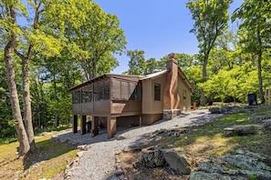A cabin with a large glass window, a brick chimney, and a gravel driveway surrounded by trees.