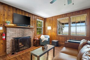 A living room with a fireplace, a television, a couch, and large windows.
