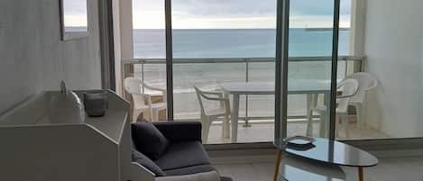 A modern living room with a sofa, a small table, and a balcony with a view of the ocean.