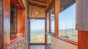 Una escalera de madera lleva a un cuarto con pared de piedra y ventanas amplias que dan a un paisaje montañoso.