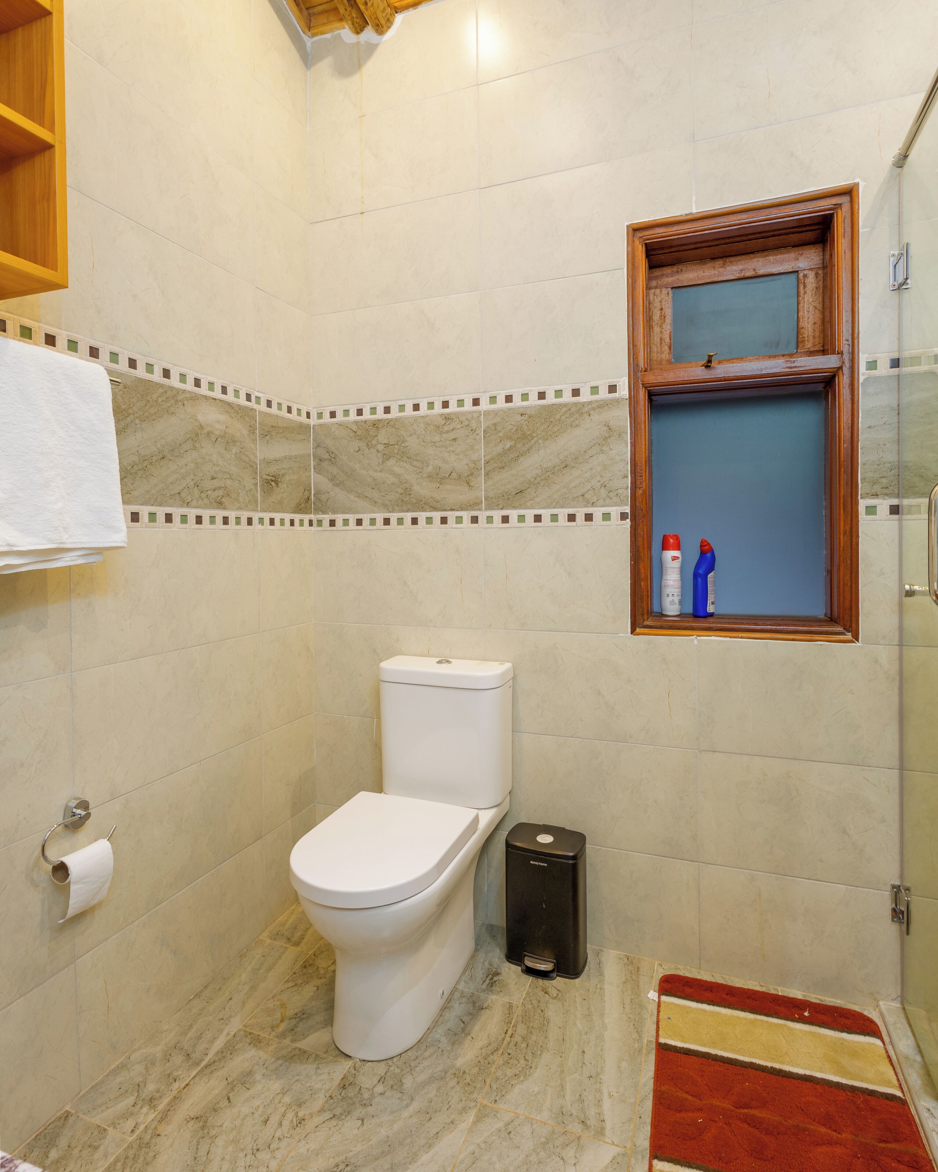 Un baño con inodoro, un cesto de basura, toallero y una ventana con productos de limpieza.