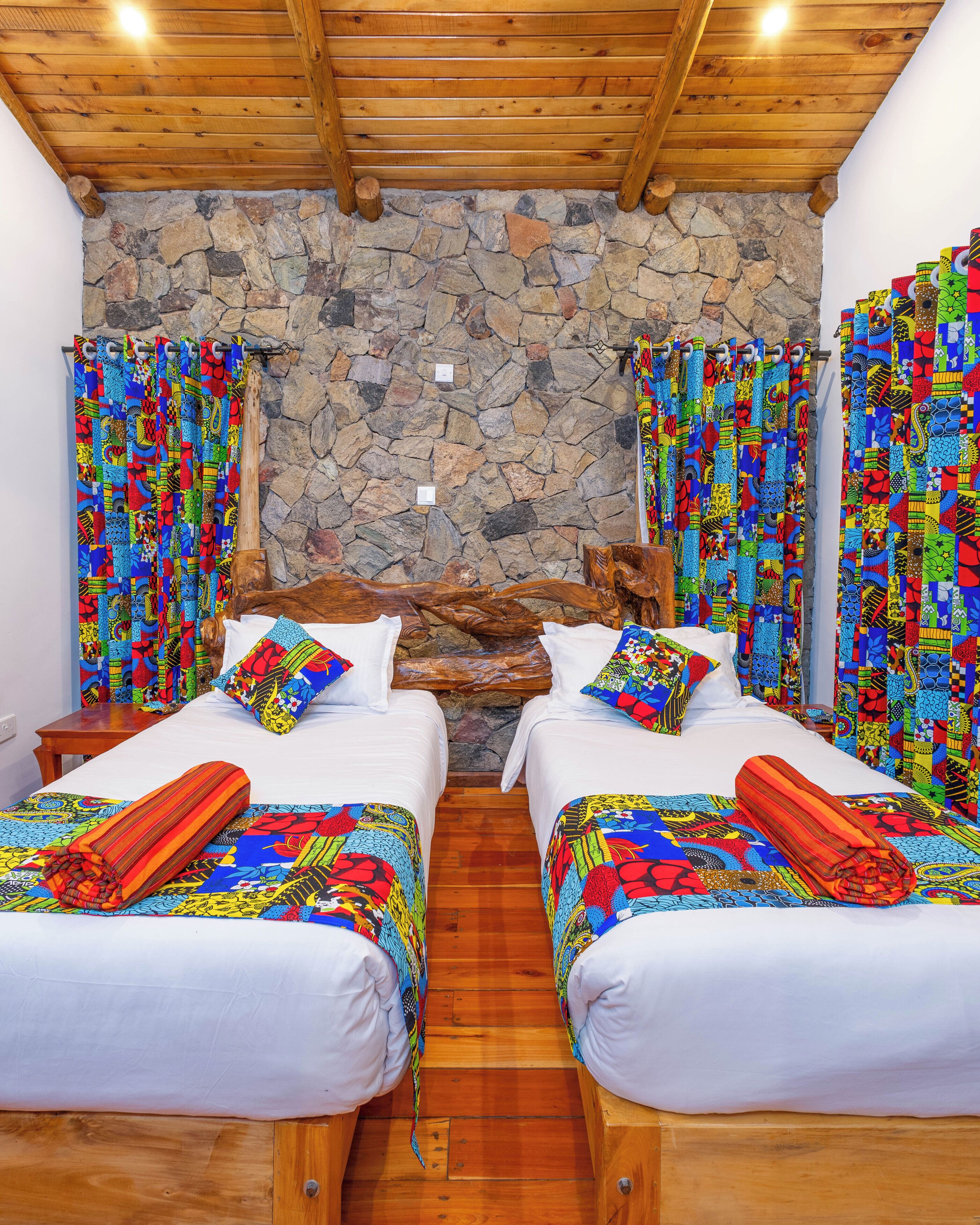 Une chambre avec deux lits, un mur en pierre, des poutres apparentes en bois au plafond et des rideaux colorés.