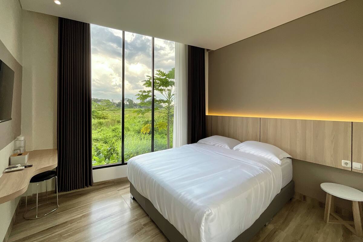 Habitación de hotel moderna con una cama grande, un escritorio y vistas a la vegetación a través de amplios ventanales.