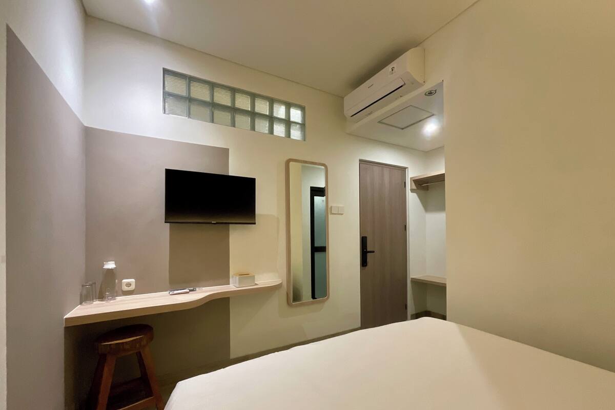Habitación de hotel con cama, televisión montada en la pared, espejo, equipo de aire acondicionado y un pequeño taburete.
