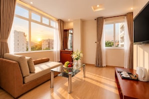 A modern living room with a sofa, a glass coffee table with fruit, a television, and a view of buildings through large windows. - G8 Diamond Hotel Saigon (Ho Chi Minh City)
