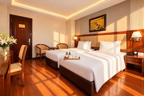 A hotel room with two beds, a desk, a chair, a nightstand, a lamp, a painting, and a vase with flowers. - G8 Diamond Hotel Saigon (Ho Chi Minh City)