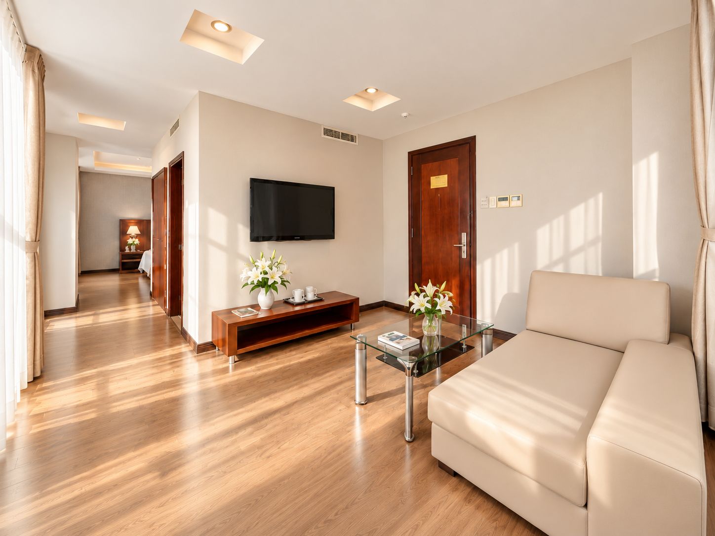 Una habitación de hotel moderna con un sofá, una mesa de centro de vidrio, un televisor montado en la pared, un mueble de entretenimiento de madera y una puerta de madera.