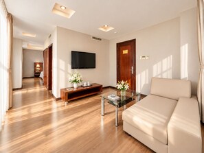 Een moderne hotelkamer met een bank, een glazen salontafel, een aan de muur bevestigde televisie, een houten entertainmentkast en een houten deur.