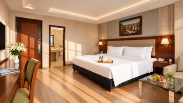 A hotel room with a large bed, bedside tables, a desk with a chair, a glass coffee table, and a painting on the wall.