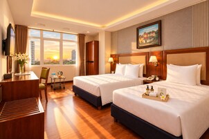 Quarto de hotel com duas camas, uma escrivaninha, uma cadeira, uma mesinha com um vaso de flores e uma janela com vista para a cidade.