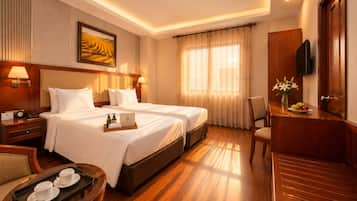 A hotel room with two beds, a desk with a chair, a TV, and a dining table with fruit.