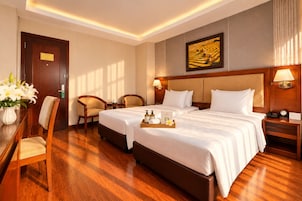 A hotel room with two beds, a desk, chairs, a painting, and a nightstand with a lamp.