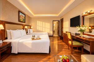 A hotel room with two beds, a desk with a chair, a television, and a dining table with fruit.