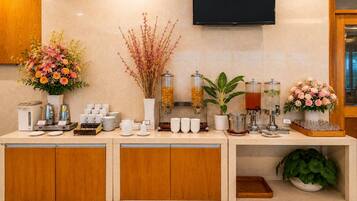 Zona de recepción de un hotel con una estación de café, un televisor montado en la pared y una variedad de flores y plantas.