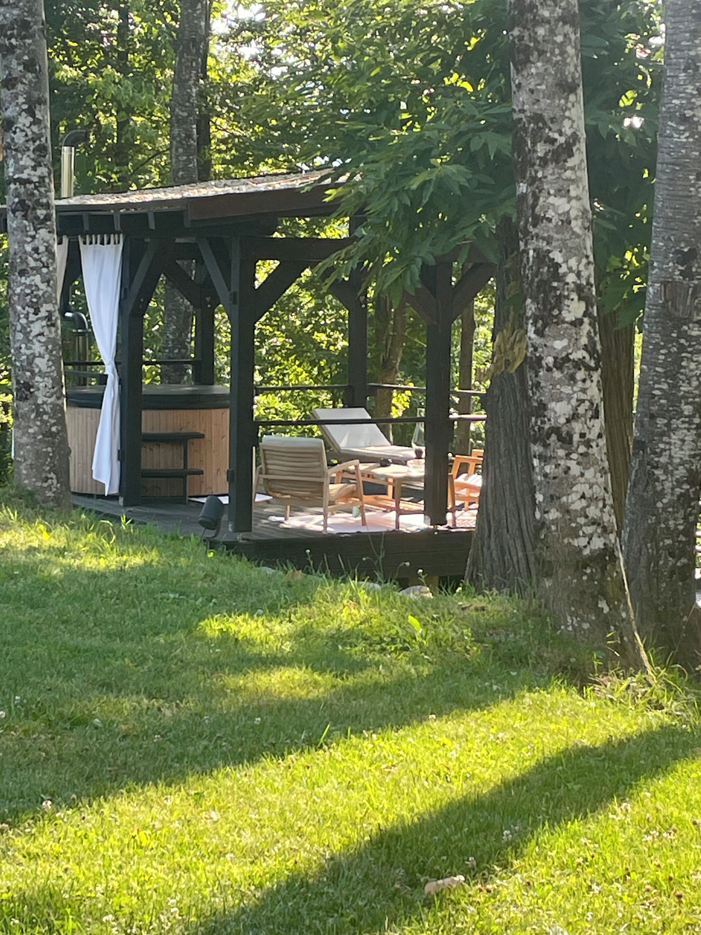 En træpavillon med bænk og bord, omgivet af træer og græs.