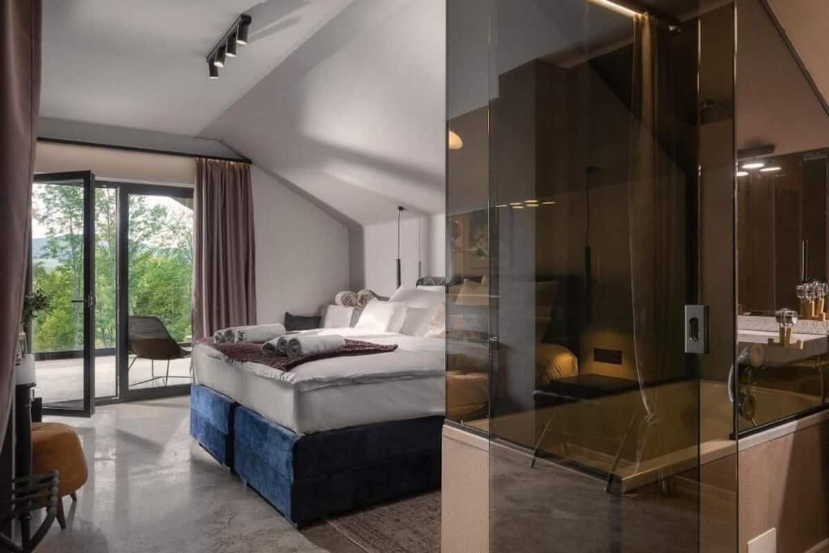 A modern bedroom with a large bed, a glass shower enclosure, and a view of greenery outside.