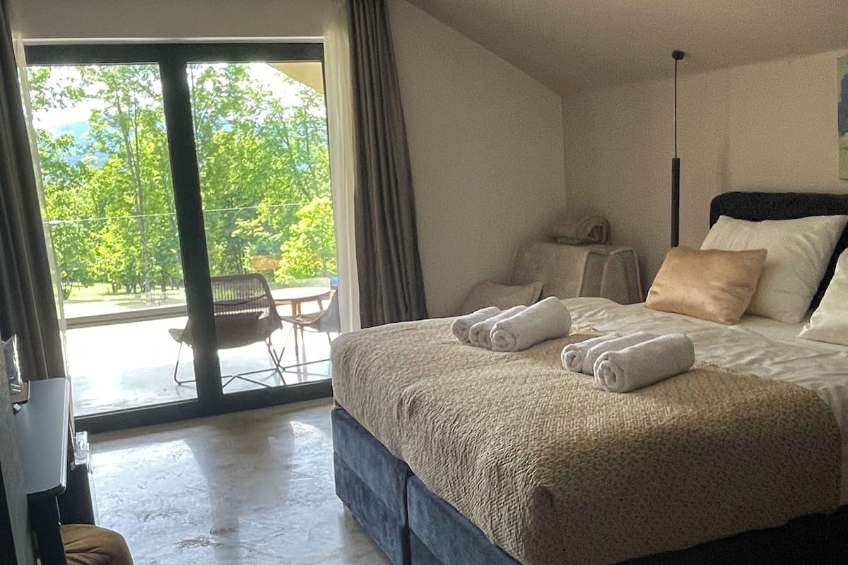 A bedroom with a large bed, a chair, a desk, and a view of greenery outside.