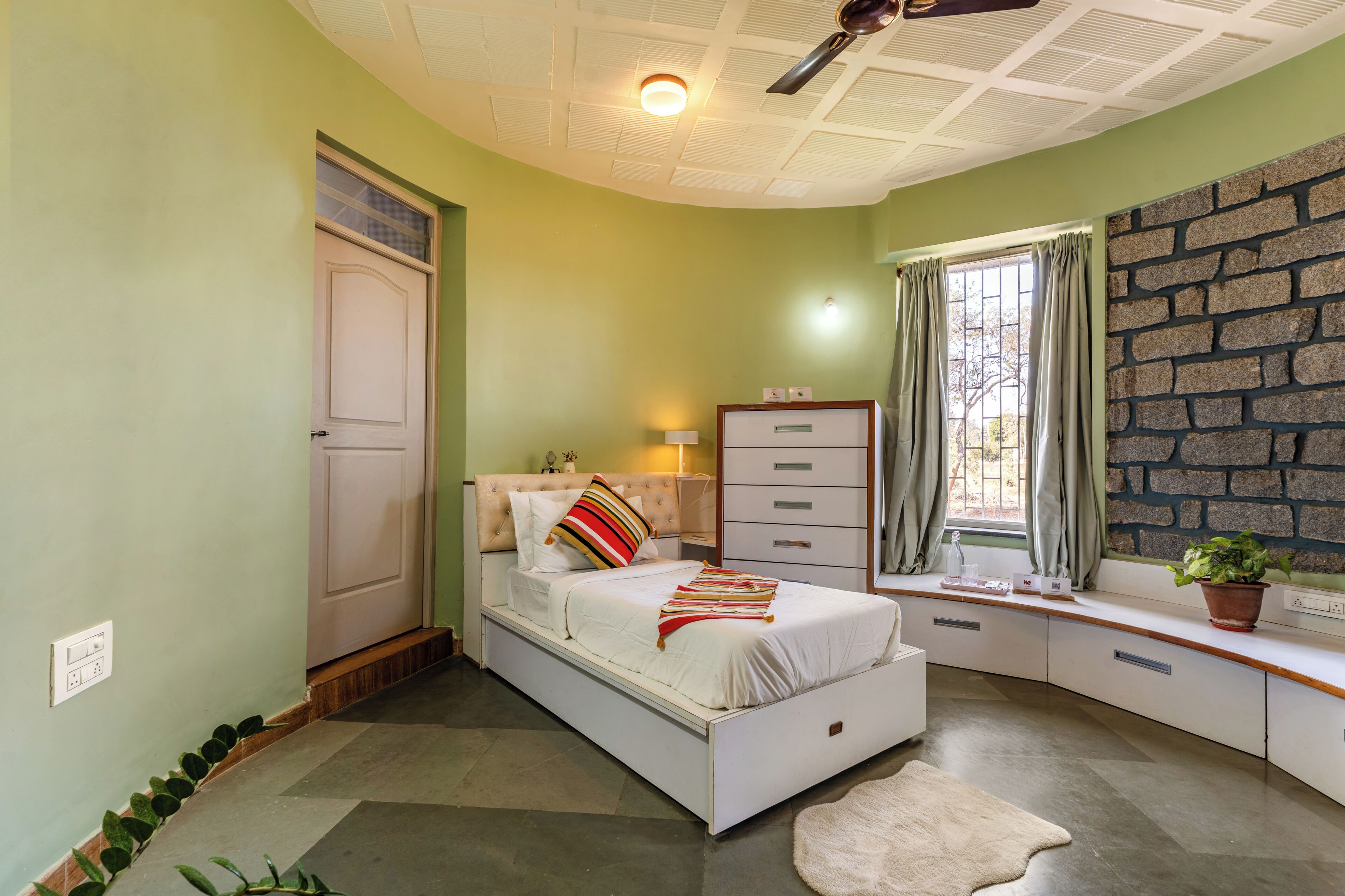 A bedroom with a bed, a window with curtains, a bedside table, a dresser, and a ceiling fan.