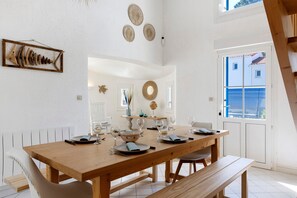 Un comedor con una mesa de madera dispuesta para cuatro personas, sillas blancas y un banco. Hay adornos colgando en la pared y se ve el exterior a través de una puerta.