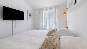 Valoinen huone, jossa on sänky, litteänäyttöinen televisio, seinään kiinnitetty lamppu ja ikkuna, jossa on läpikuultavat verhot.