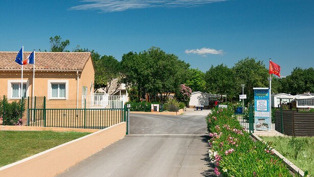 A road leading to a building with flags, a signboard, and a fenced area with flowers. - Mobilhome in a campsite with swimming pool. Close to the sea. Pets allowed. (Saint-Thibéry)
