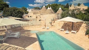 A pool area with lounge chairs, umbrellas, and stone buildings in the background. - Navolte Trulli e Piscina (Locorotondo)