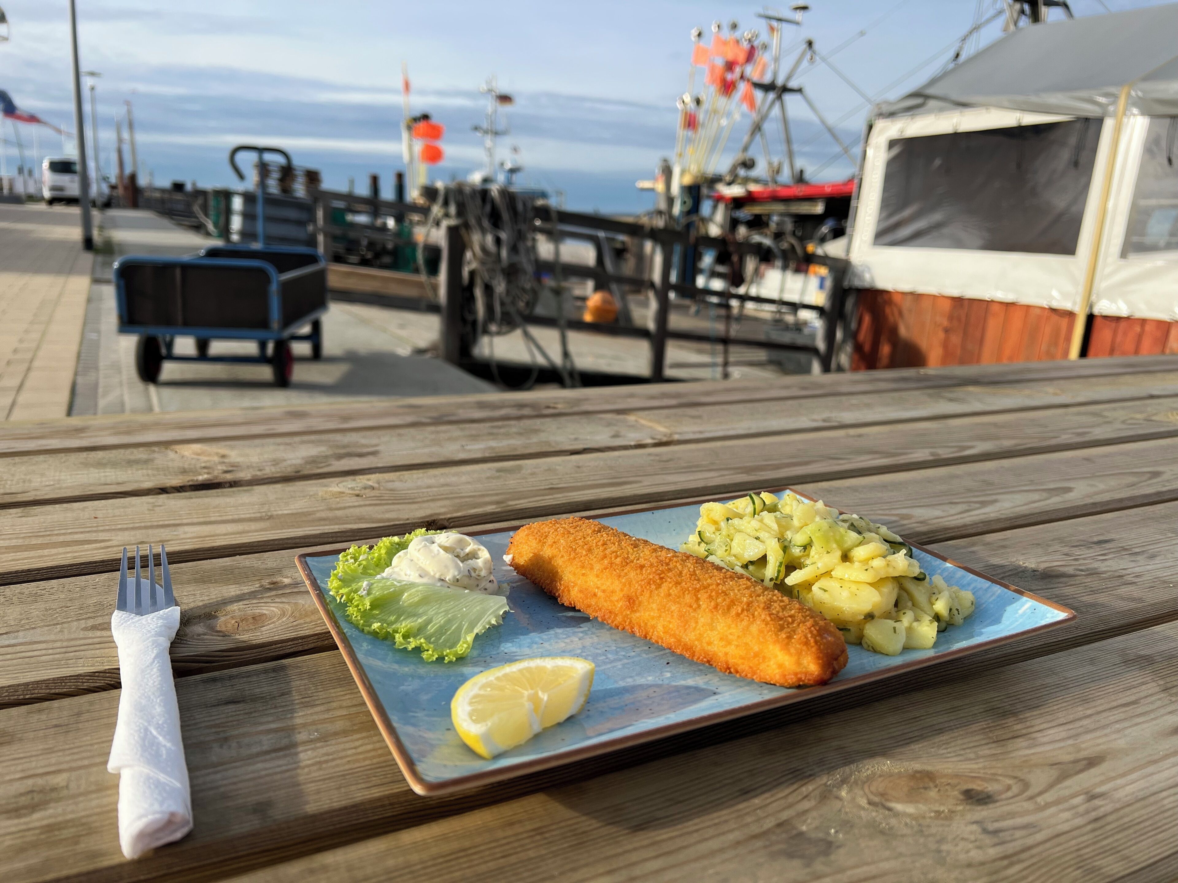 A meal on a wooden table with a fork and napkin, a plate with a breaded item, potatoes, and a side salad.
