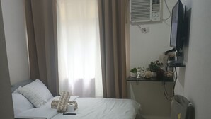 A hotel room with a bed, a desk, a chair, a television, an air conditioning unit, and a window with curtains.