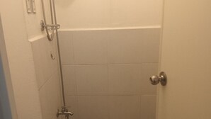 A small, narrow bathroom with a toilet, a shower, and a bucket.