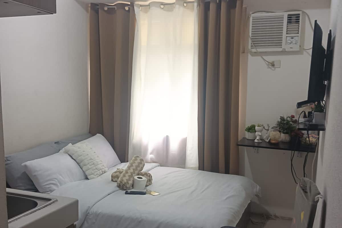 A bedroom with a bed, a desk, a chair, a TV, and an air conditioning unit.