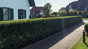 Ein Weg führt zu einem weißen Gebäude mit grünen Fensterläden, umgeben von Hecken und weiteren Häusern.