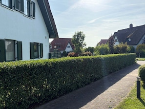 Ein Weg führt zu einem weißen Gebäude mit grünen Fensterläden, umgeben von Hecken und weiteren Häusern.