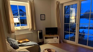Ein Wohnzimmer mit einer Couch, einem Fernseher, einem kleinen Tisch mit einer Schale und Blick auf eine verschneite Landschaft durch eine Glastür.
