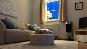 Ein Wohnzimmer mit einer Couch, einem Hocker, einer Lampe, einem Fernseher und einem Fenster mit Vorhängen.