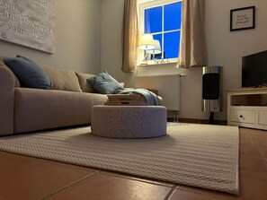 Ein Wohnzimmer mit einer Couch, einem Hocker, einer Lampe, einem Fernseher und einem Fenster mit Vorhängen.