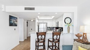 A modern kitchen with white cabinets, a stainless steel refrigerator, and a microwave. There are two wicker chairs at a small island with a sink. - Enjoy stunning ocean views from the balcony, living room, dining area, and kitchen. This 2BR condo features a heated beachfront pool, fitness room, king beds in both bedrooms, and comfortably sleeps 4 in beautiful Daytona Shores. (Daytona Shores)