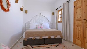 Un dormitorio con cama, una puerta de madera, una ventana con cortinas y un espejo fijado a la pared.