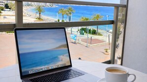 Un ordinateur portable est posé sur une table de balcon, à côté d’une tasse de café, avec vue sur une plage bordée de palmiers et un ciel dégagé.
