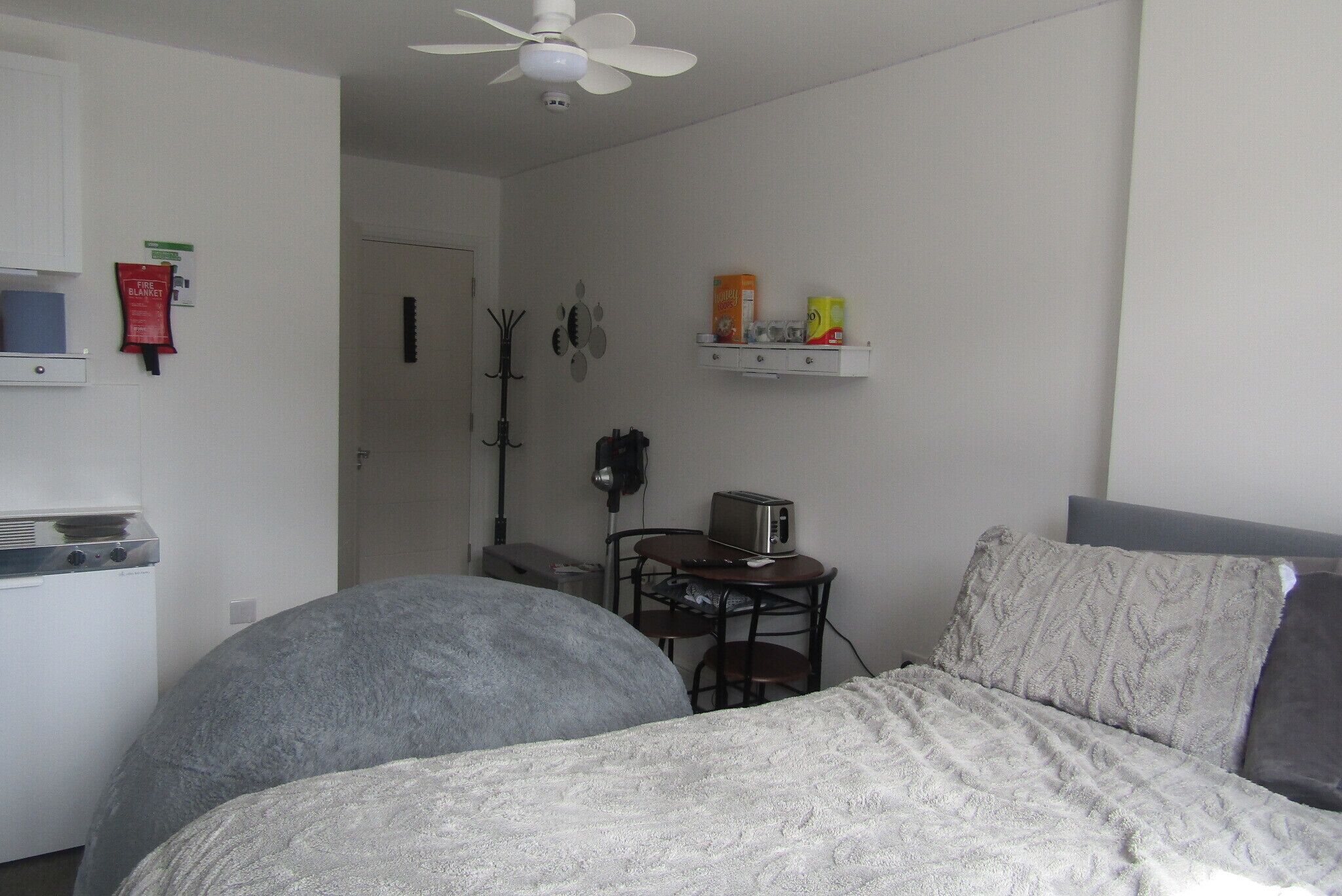 A bedroom with a bed, a ceiling fan, a small table with a toaster, and a shelf with items.
