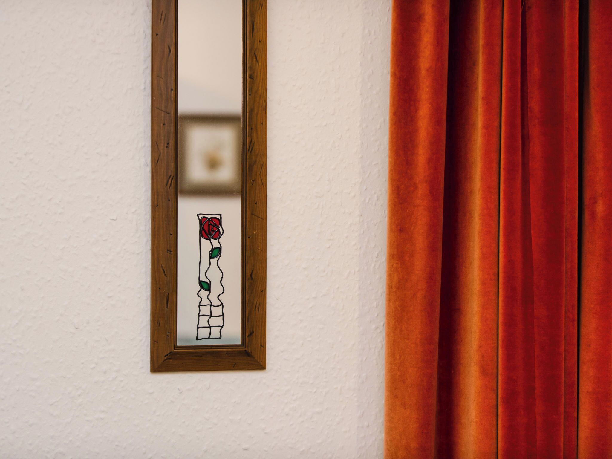 A mirror reflecting a red rose and a white vase on a white wall.