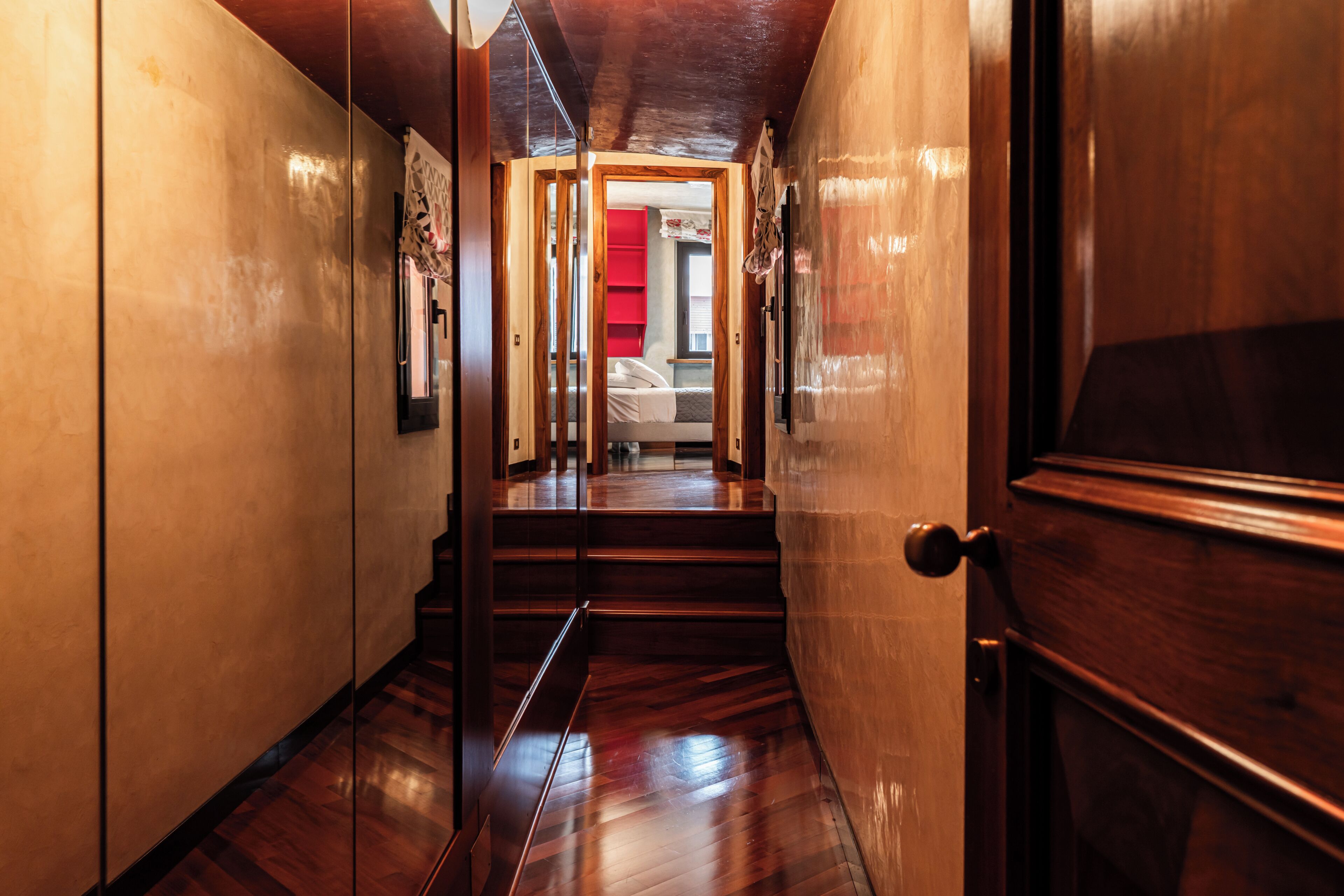 A hallway with wooden floors and walls, leading to a room with a bed and a red cabinet.
