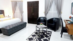 Quarto de hotel com cama, sofá, duas poltronas, uma mesinha com um vaso, escrivaninha com um laptop e uma porta grande.